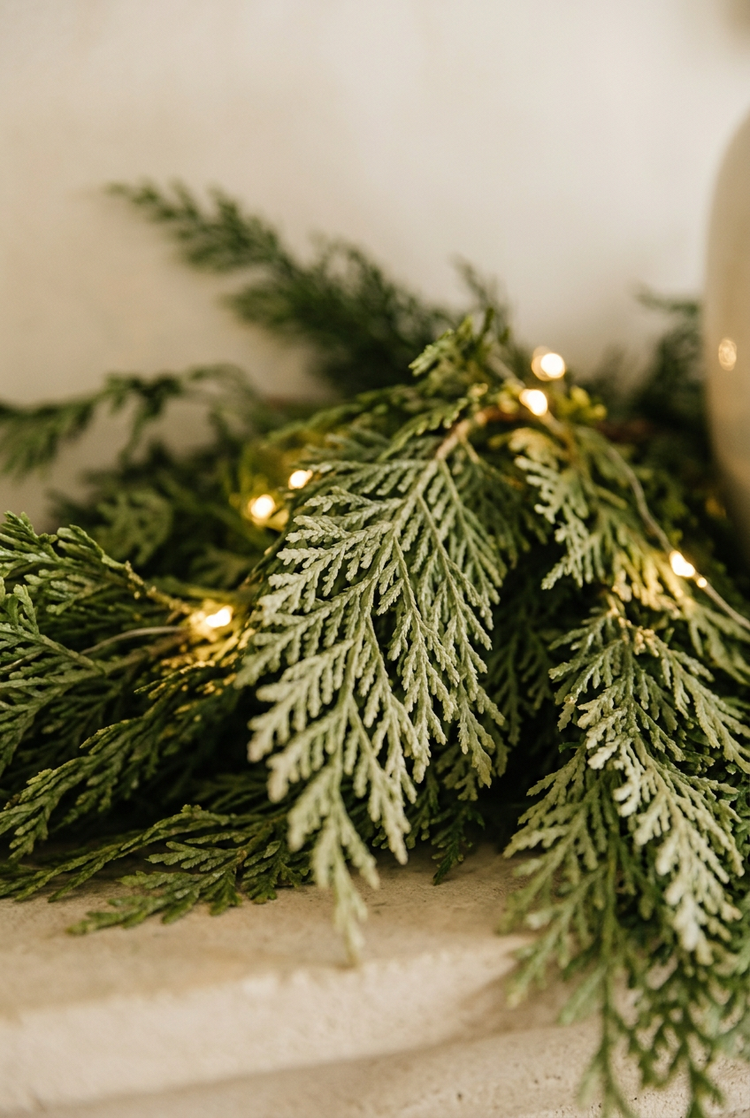Close-up of soft faux cedar garland with warm white lights on a cream mantel